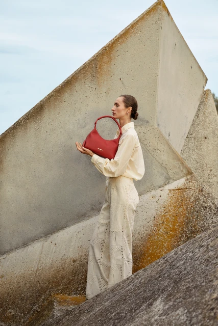 Woman in a cream-colored outfit poses with a red leather handbag in front of modern concrete blocks, showcasing fashion and design.