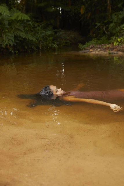 An adult female floats in a serene river surrounded by lush green trees, creating a tranquil nature scene with warm, natural light.