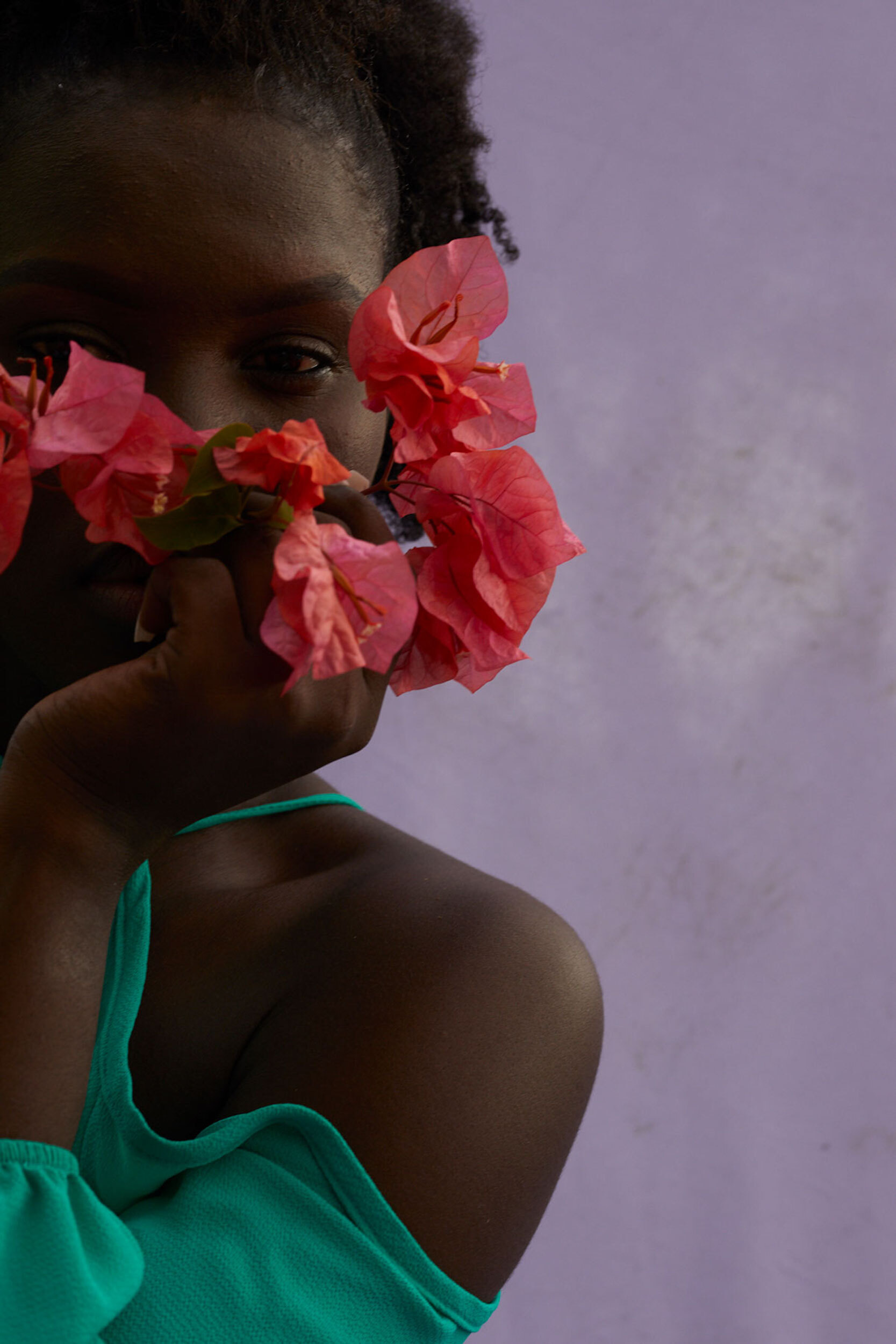 Woman holds bougainvillea flowers, partially covering face, wearing turquoise top in a portrait against a light purple background.