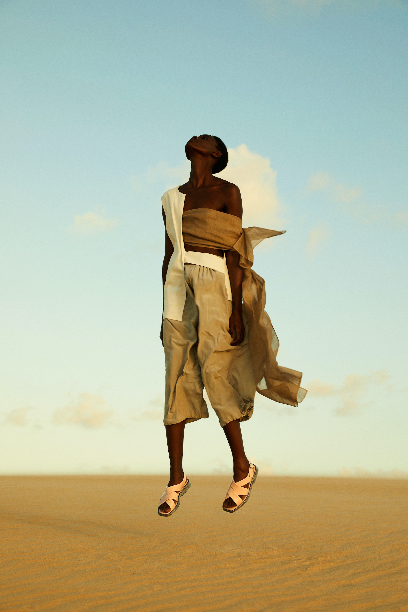 Woman in flowing beige and white outfit levitates in a desert landscape, sandy ground and bright blue sky with light clouds.