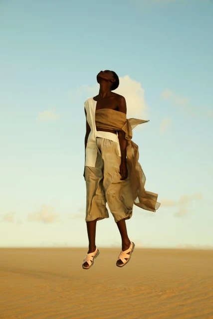 Woman in flowing beige and white outfit levitates in a desert landscape, sandy ground and bright blue sky with light clouds.