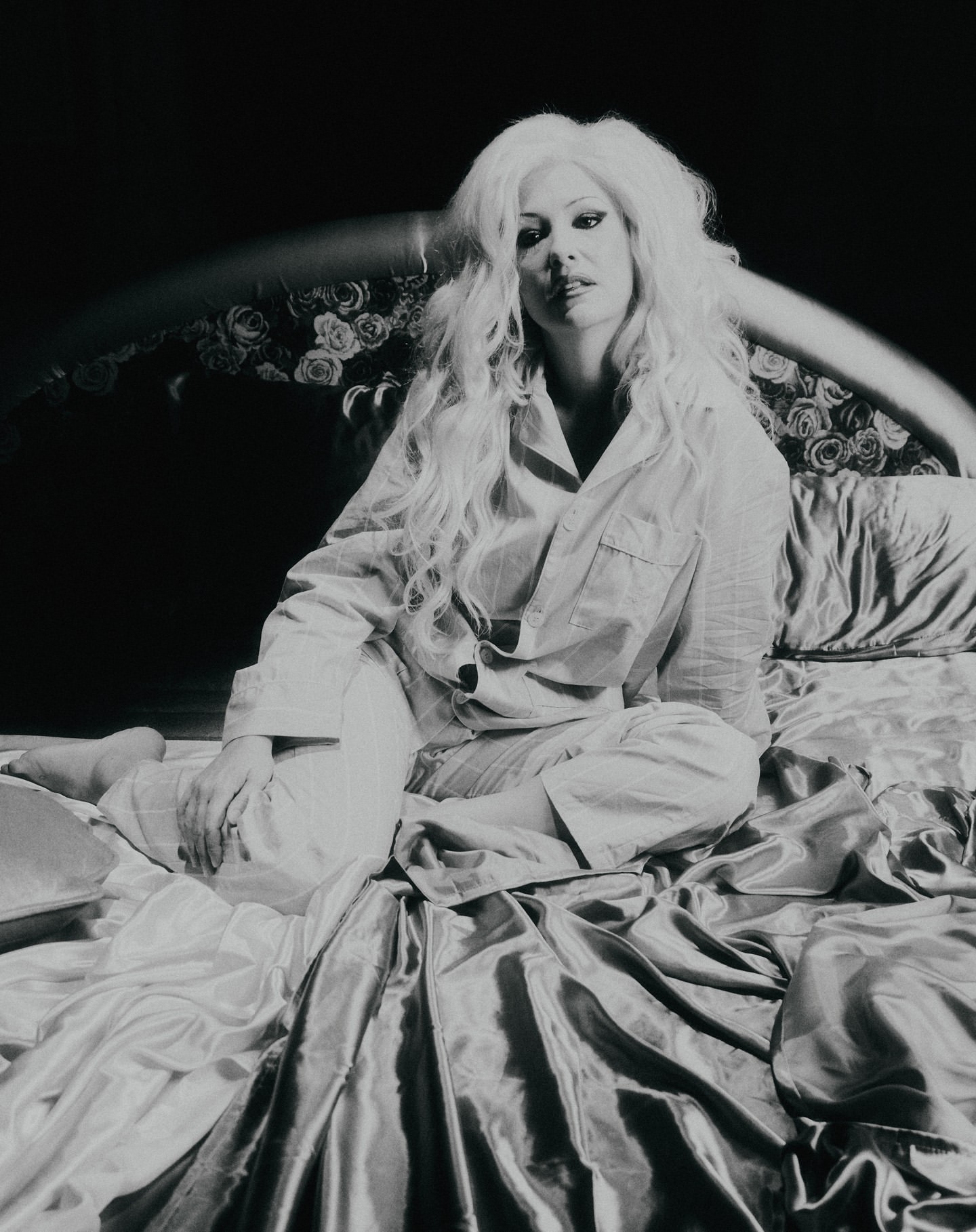 Black and white portrait of a woman with long wavy hair in pajamas, sitting on a satin bed with a floral headboard.