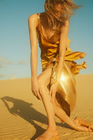 Model in a shiny gold dress standing on sand dunes with long windblown blonde hair and bright sunlight casting a shadow