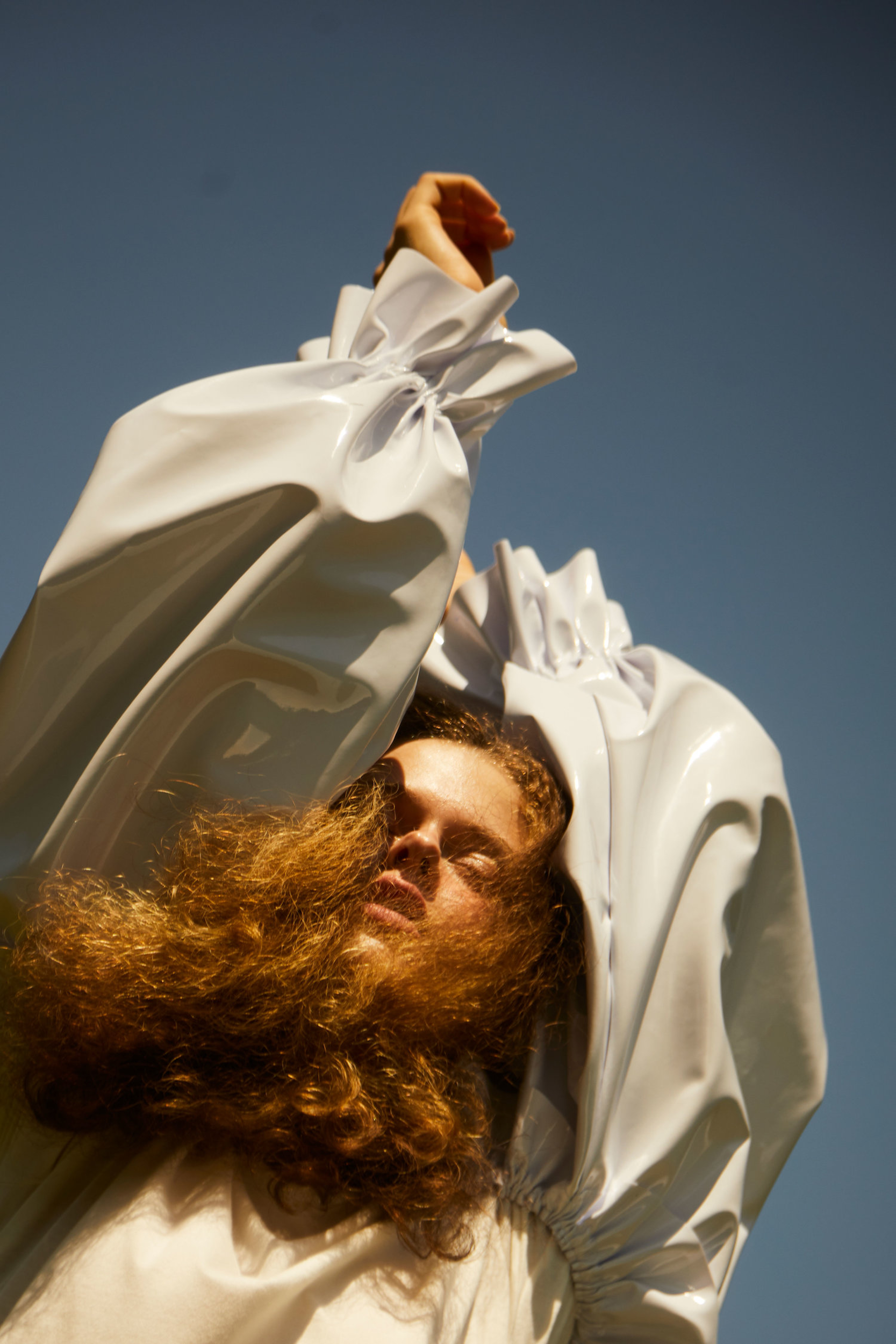 Portrait of a person with long curly hair wearing a glossy white sculptural garment against a clear blue sky.