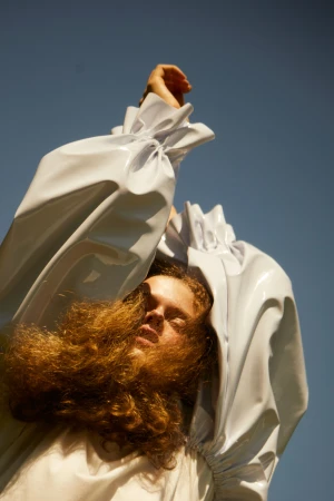 Portrait of a person with long curly hair wearing a glossy white sculptural garment against a clear blue sky.