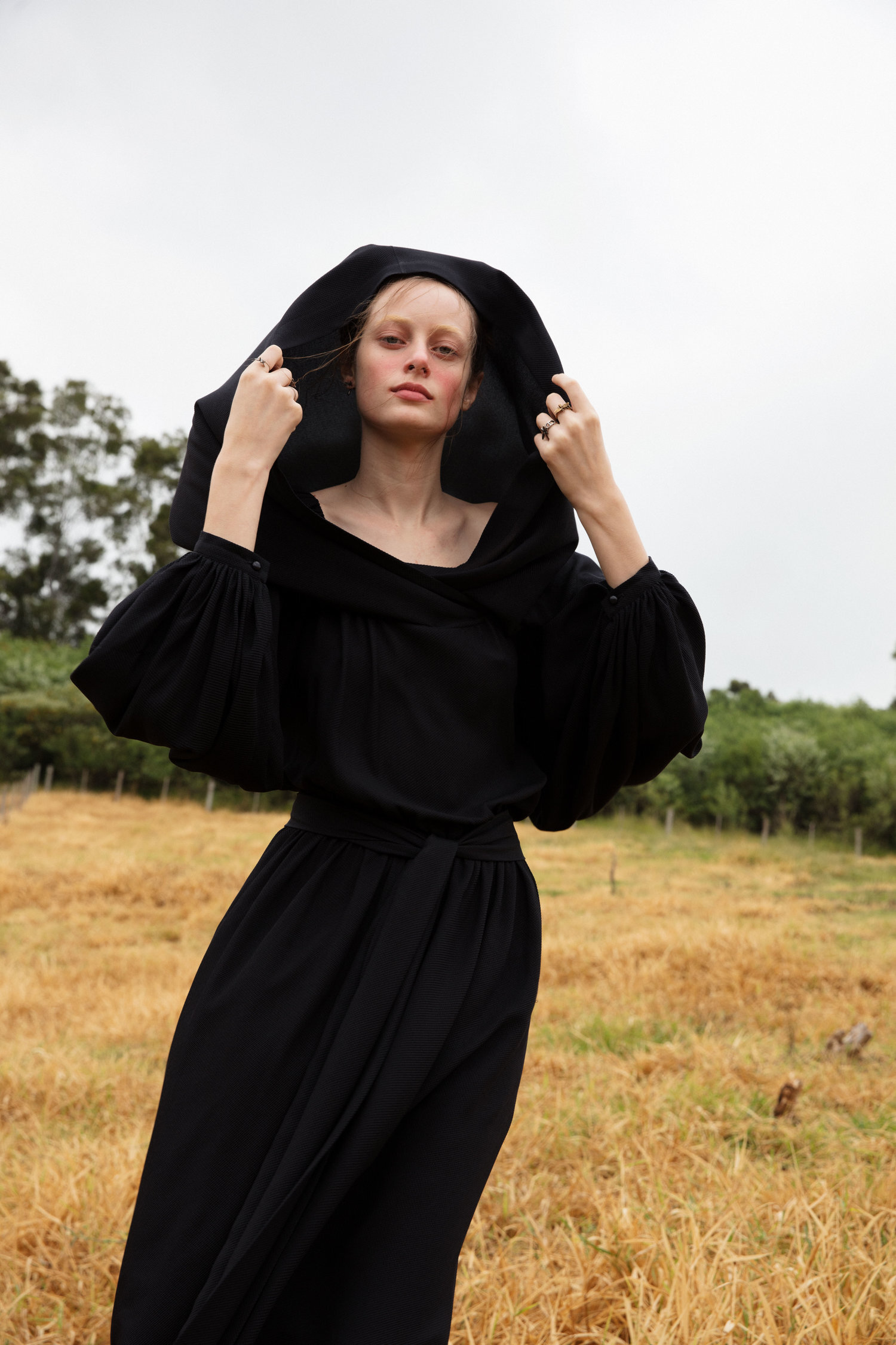 Model in a black hooded dress standing in a dry grassy field under an overcast sky, photographed by Cecilia Duarte.