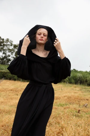 Model in a black hooded dress standing in a dry grassy field under an overcast sky, photographed by Cecilia Duarte.