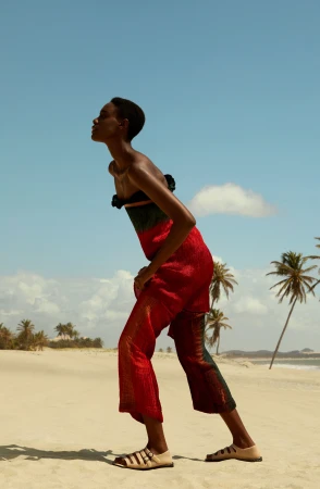 Black woman in red and green textured jumpsuit posing on a sandy beach with palm trees under a clear blue sky