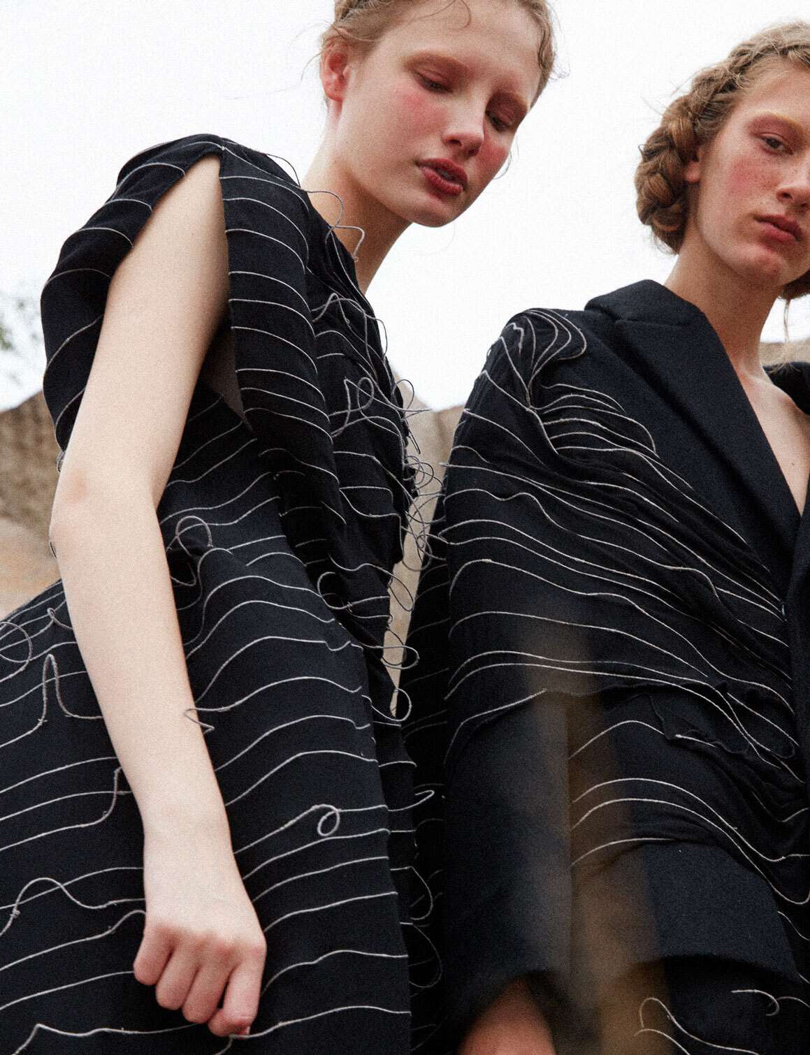 Two women in black dresses with white line designs pose outdoors, showcasing fashion and style in a soft, overcast light setting.
