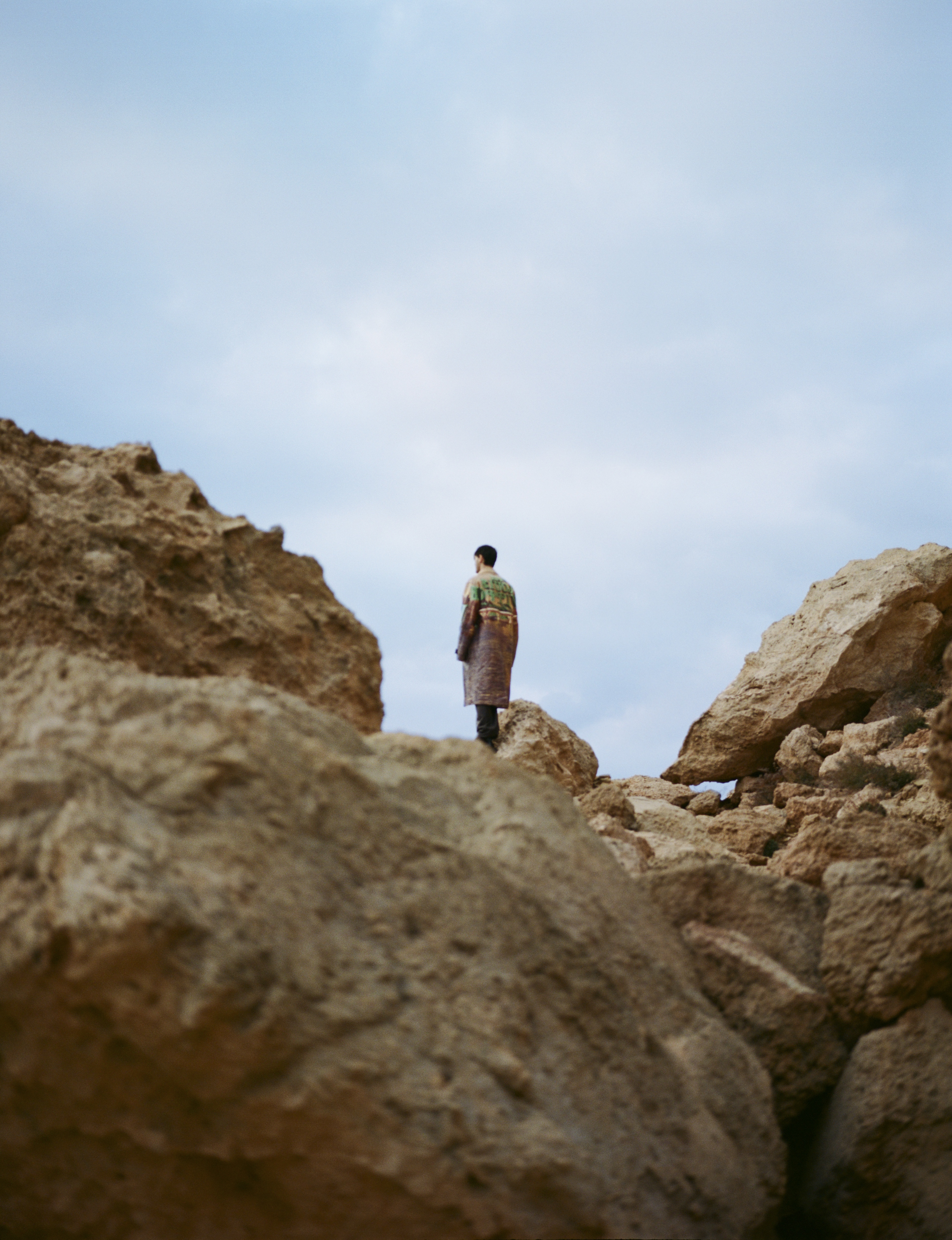 Man in patterned robe stands on rocky terrain under cloudy sky. Outdoor fashion shot, low angle, natural light, neutral colors.