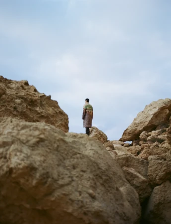 Man in patterned robe stands on rocky terrain under cloudy sky. Outdoor fashion shot, low angle, natural light, neutral colors.