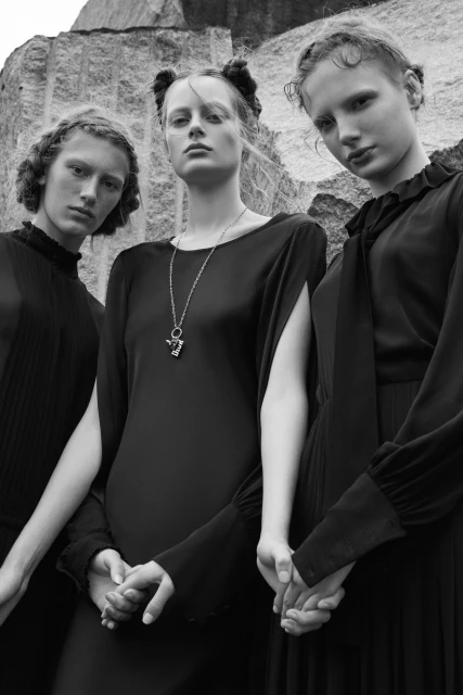 Monochrome fashion shot of three women in dark dresses against a quarry backdrop, holding hands with serious expressions.