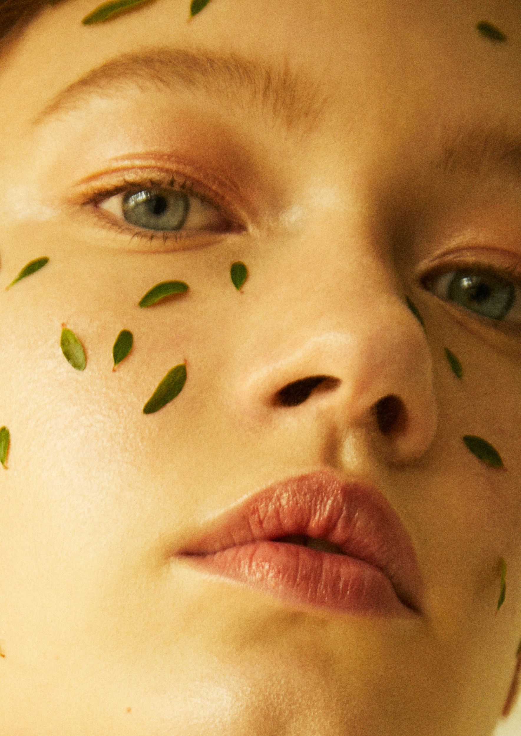 Close-up portrait of a fair-skinned adult woman with scattered green leaves on her face, styled for a beauty or skincare campaign.