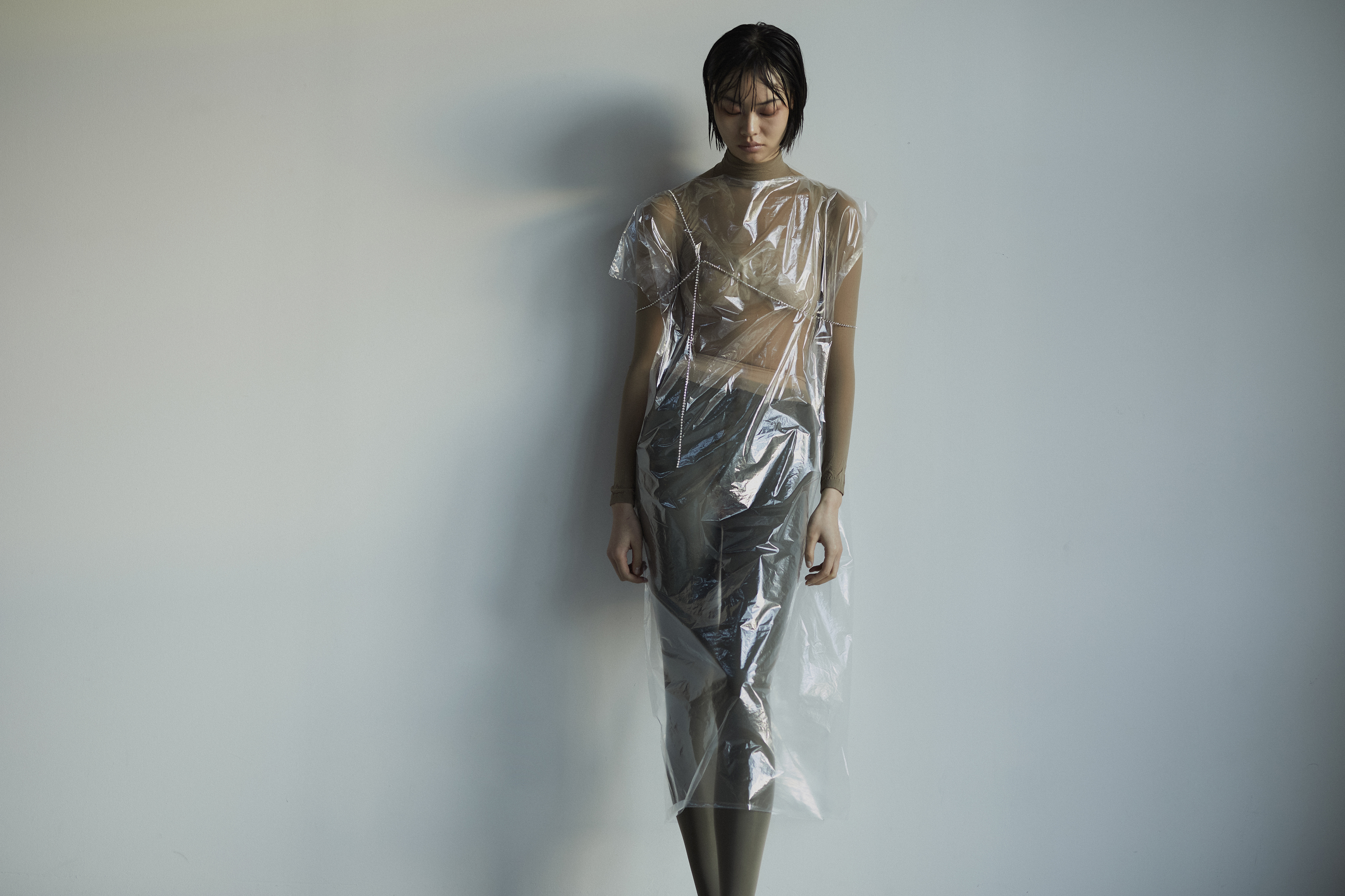 Female model posing in a translucent plastic dress against a plain wall, showcasing unique fashion and studio lighting. 