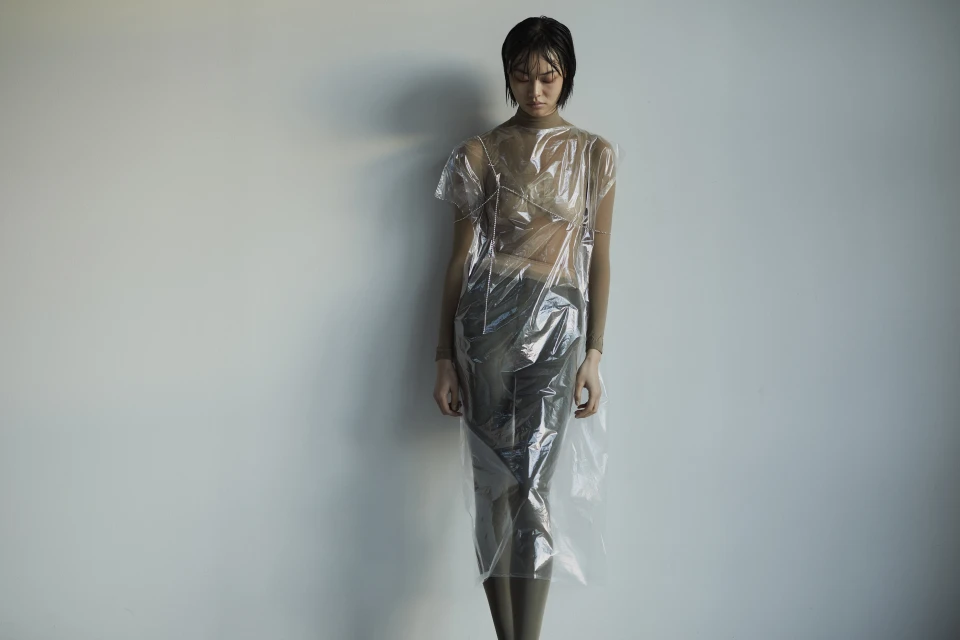 Female model posing in a translucent plastic dress against a plain wall, showcasing unique fashion and studio lighting. 