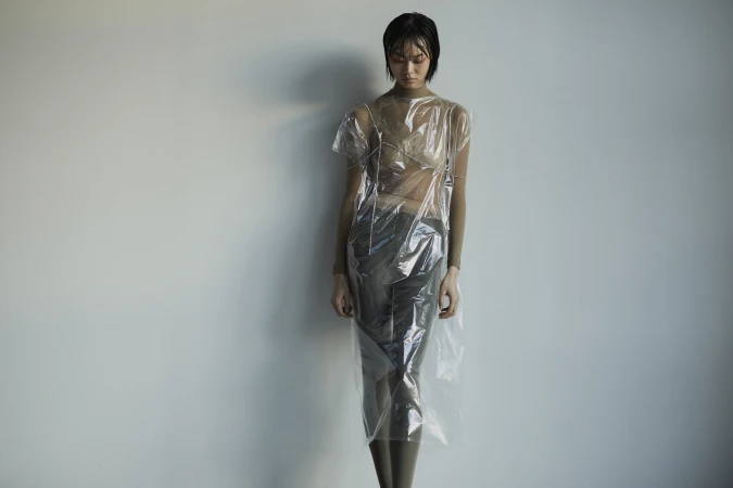 Female model posing in a translucent plastic dress against a plain wall, showcasing unique fashion and studio lighting. 