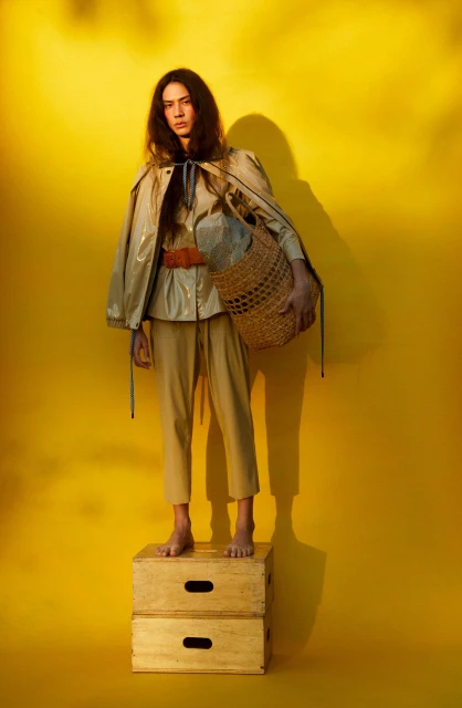 Woman in a tan outfit standing on wooden crates holding a woven bag against a vibrant yellow background, creating a striking fashion shot.