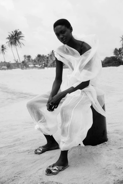 Black and white portrait of a woman in a white outfit, sitting on an object on a sandy beach with palm trees in the background.