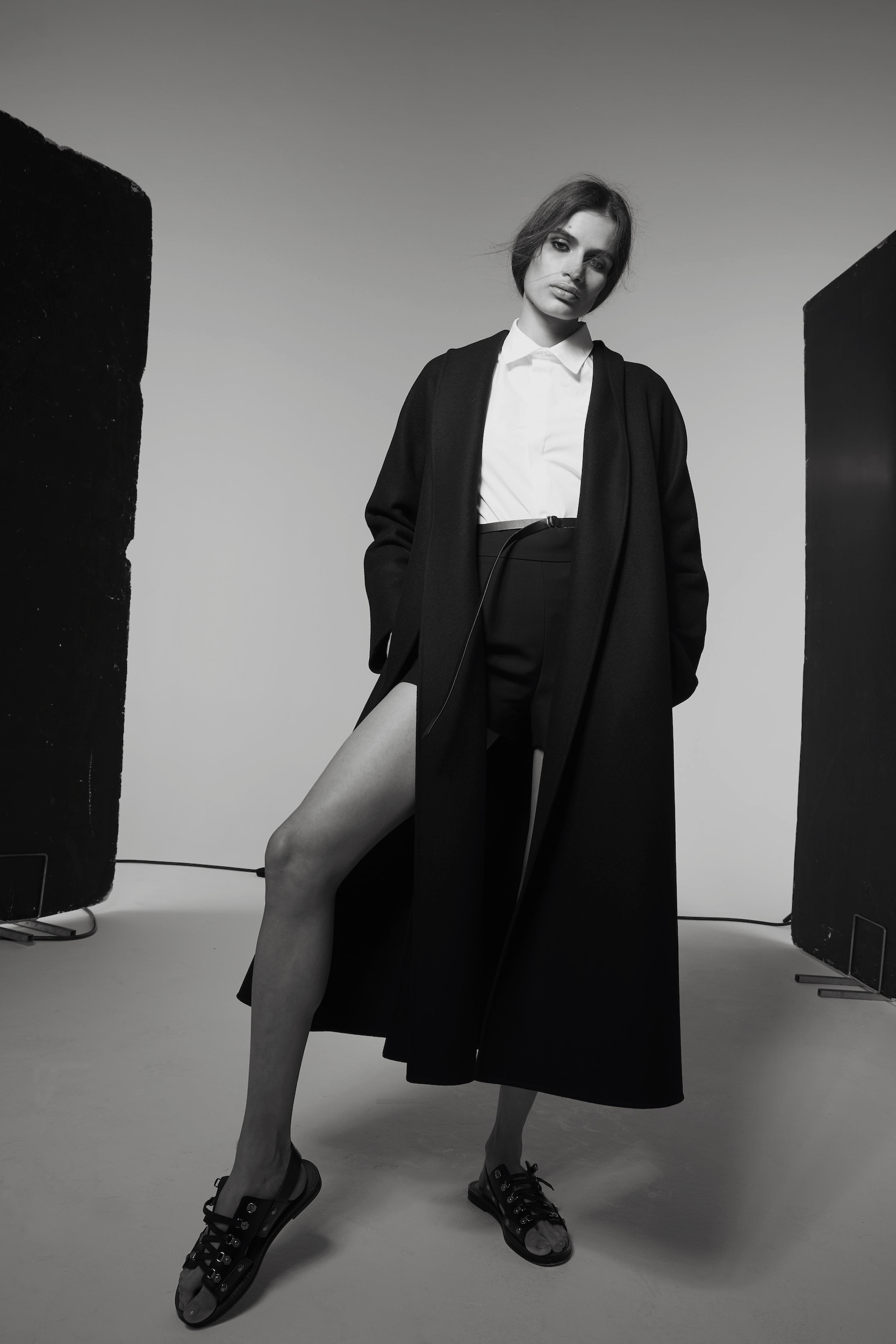 Monochrome portrait of a fashionable woman in a white top, black shorts, and a long black coat posing in a photo studio.