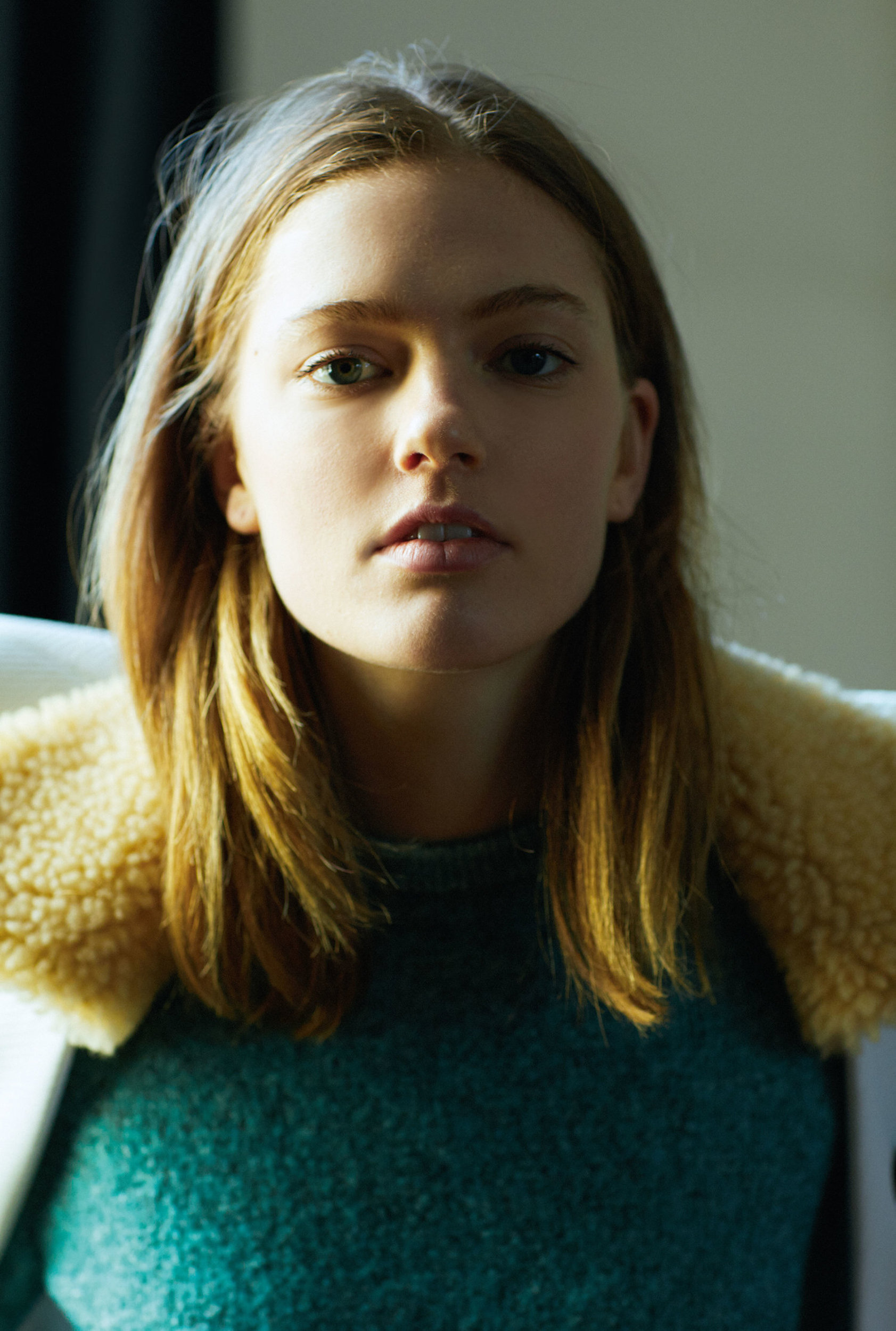 Close-up portrait of a young adult woman with long brown hair and a teal sweater, illuminated by soft, natural lighting in an indoor setting.