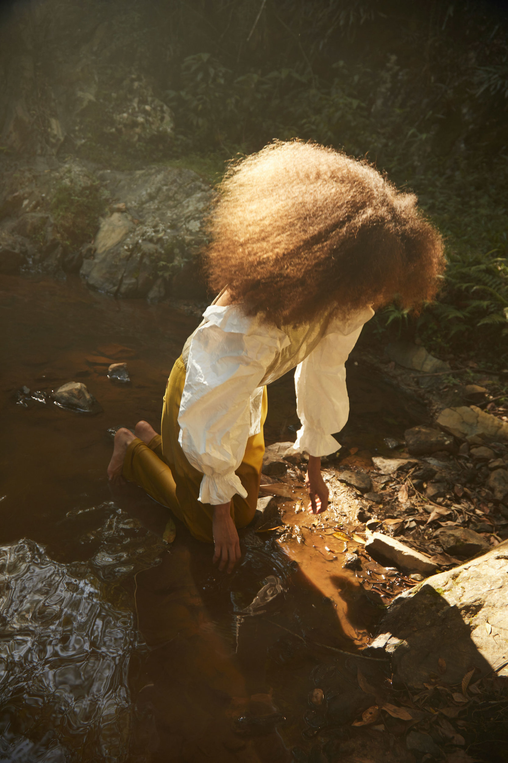 Young woman with voluminous curly hair kneeling near a shallow stream outdoors, wearing a white blouse and yellow pants, captured in soft light.
