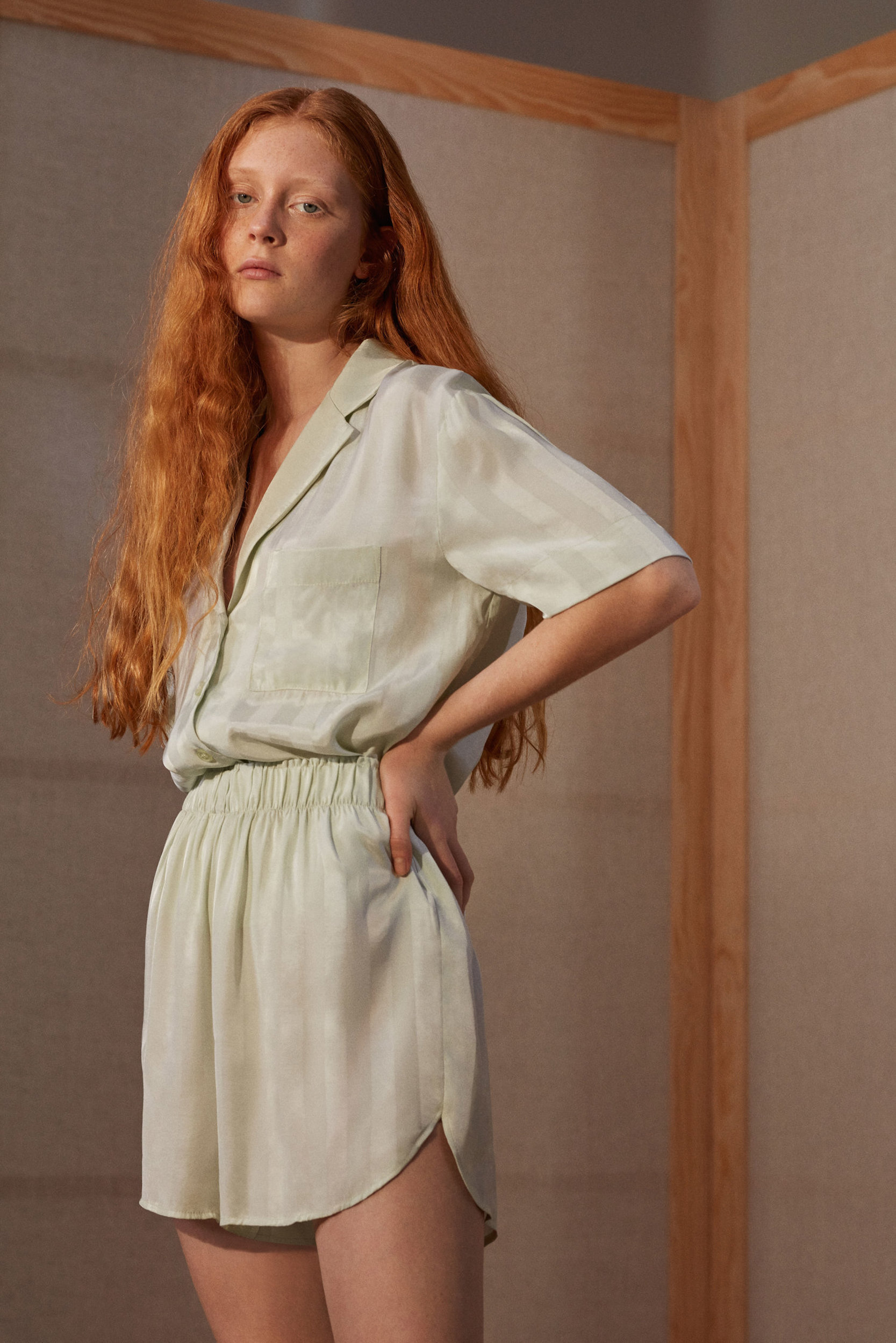 Woman with long red hair wearing matching light green lounge set with short sleeves, shorts, hands on hips in a minimalist setting.