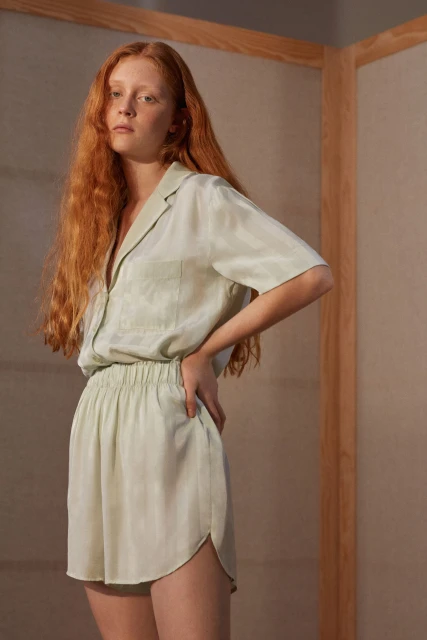 Woman with long red hair wearing matching light green lounge set with short sleeves, shorts, hands on hips in a minimalist setting.