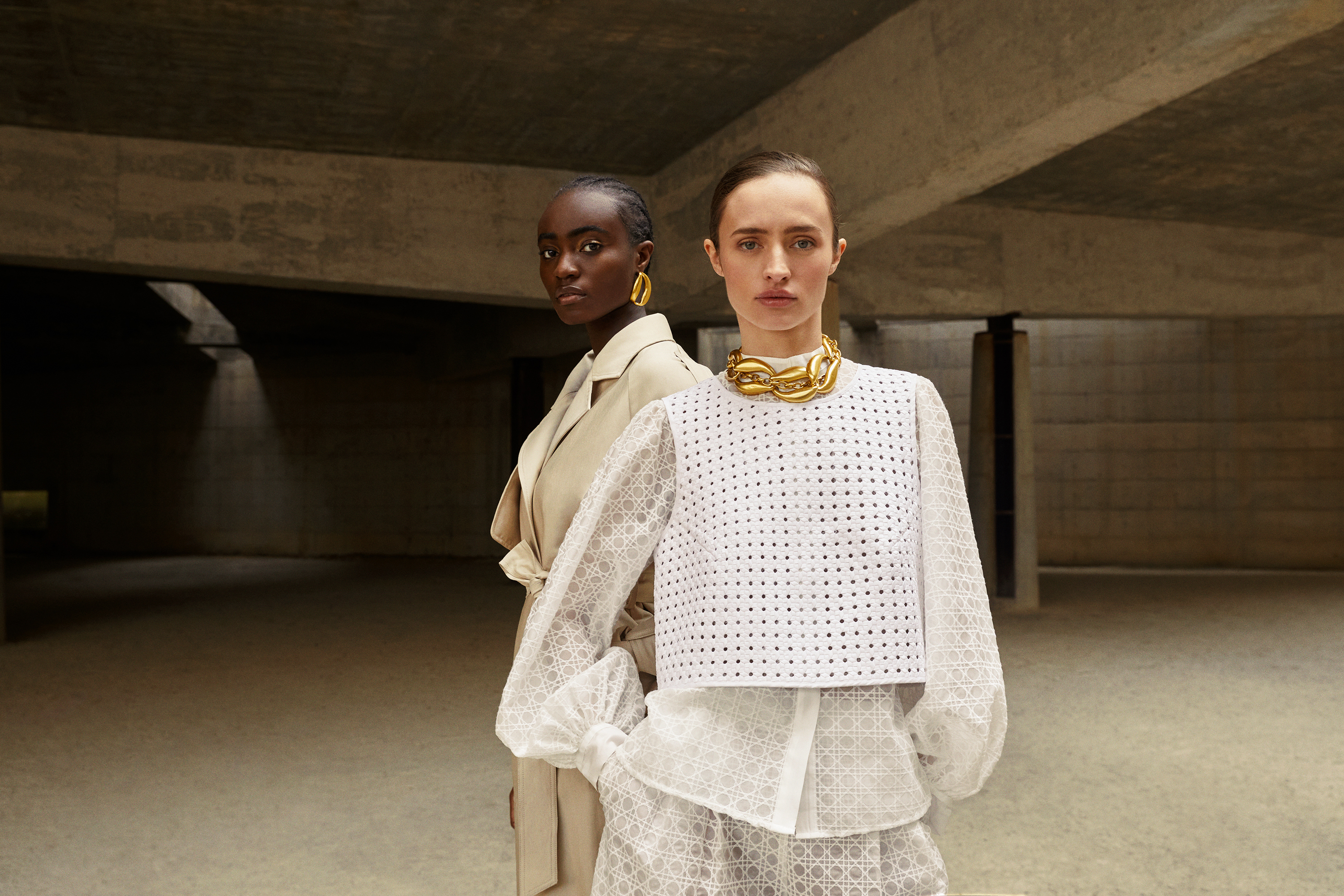 Two fashion models in a concrete setting, one wearing a beige trench coat and earrings, the other a white top and gold necklace.