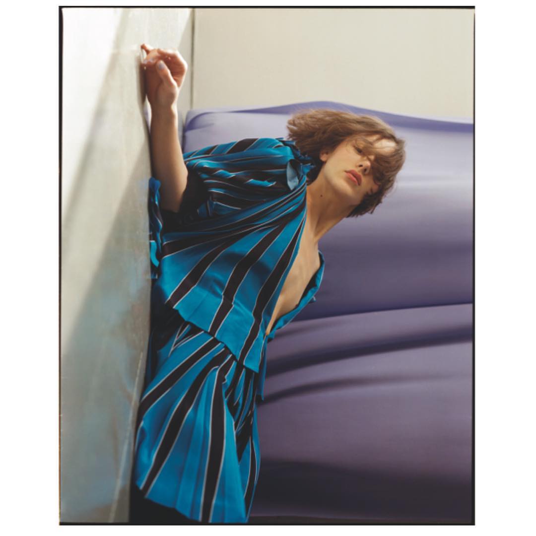 A young adult leans against a wall wearing a teal and black striped robe or shirt. The indoor scene features soft natural lighting.