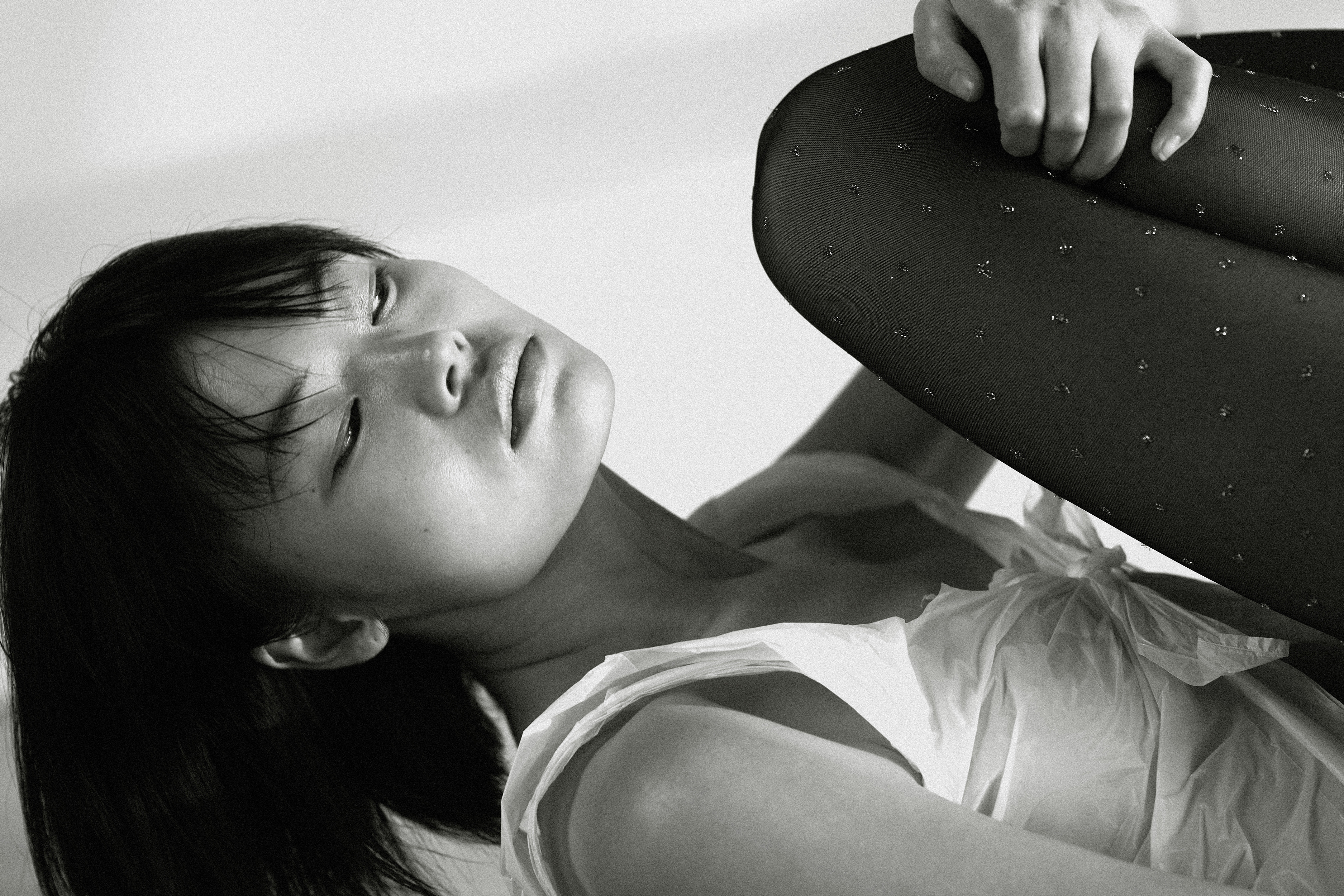 Monochrome portrait of an Asian woman in a white ruffled top and dotted black tights, resting her head with eyes closed on a white backdrop.