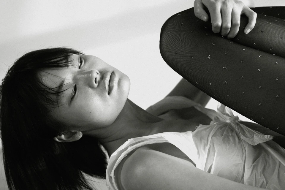 Monochrome portrait of an Asian woman in a white ruffled top and dotted black tights, resting her head with eyes closed on a white backdrop.