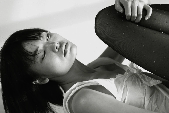 Monochrome portrait of an Asian woman in a white ruffled top and dotted black tights, resting her head with eyes closed on a white backdrop.