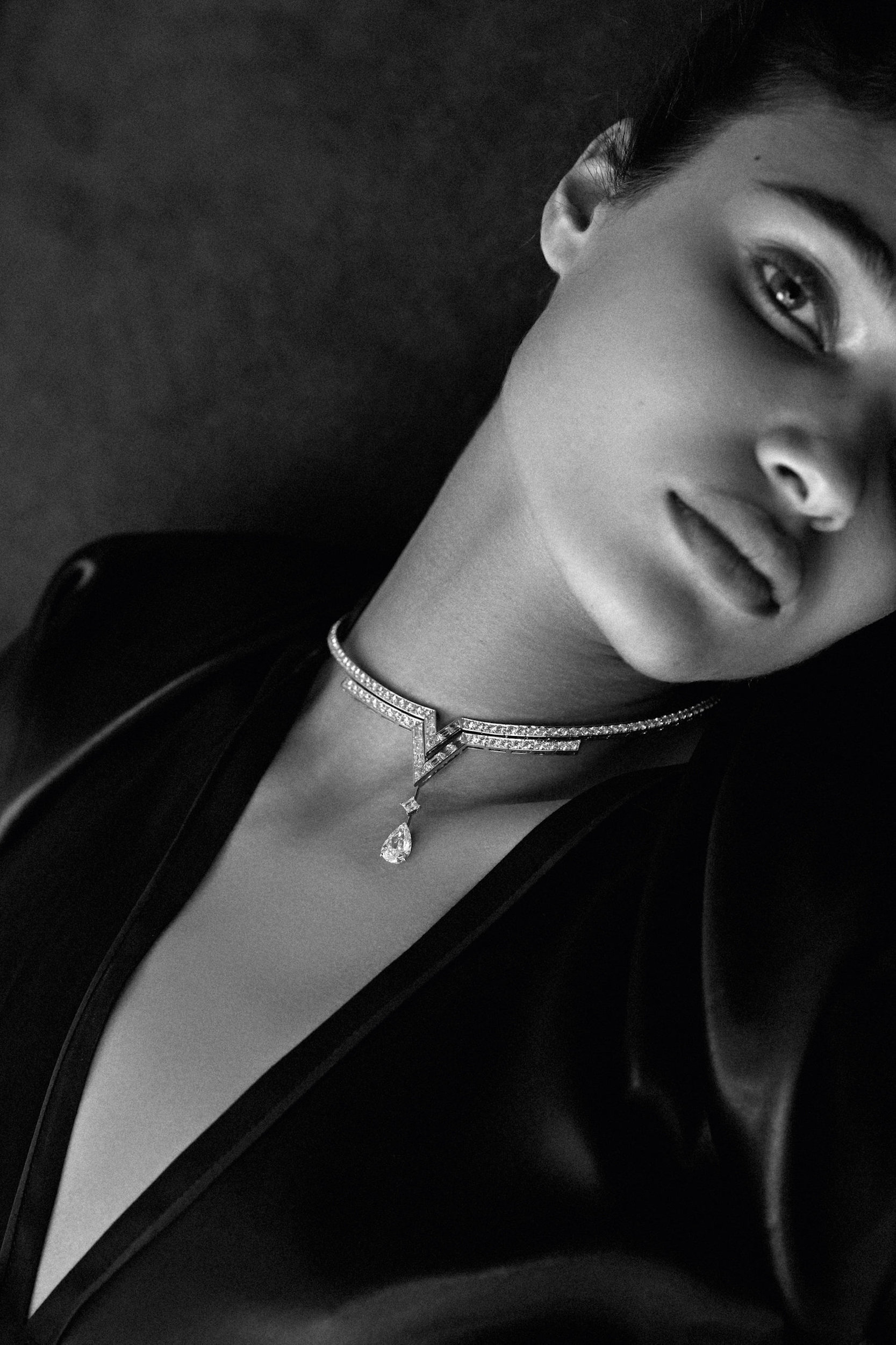 Monochrome portrait of a woman wearing a black top and a delicate diamond choker necklace against a dark grey backdrop.