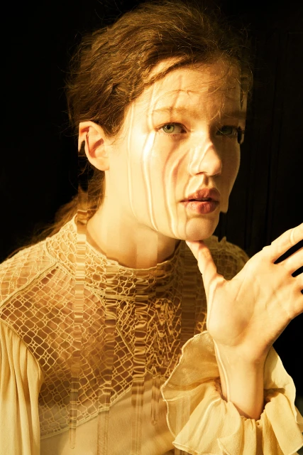 Woman with wavy hair in patterned dress, hand near face, bathed in warm light against a dark background. Beauty and fashion shot.