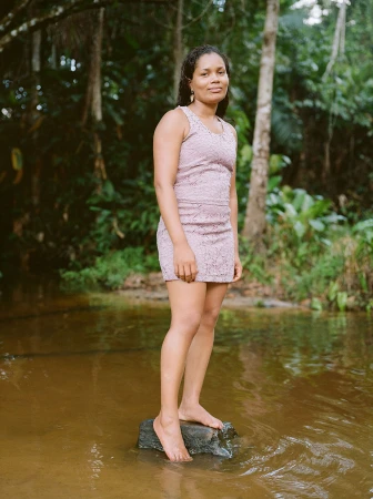 Woman in a pink lace mini dress standing barefoot on a rock in a shallow river surrounded by a dense forest.