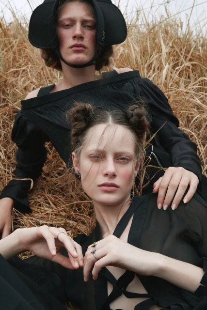 Two fair-skinned fashion models pose in tall grass wearing black outfits and headwear, showcasing a blend of high fashion and natural setting.
