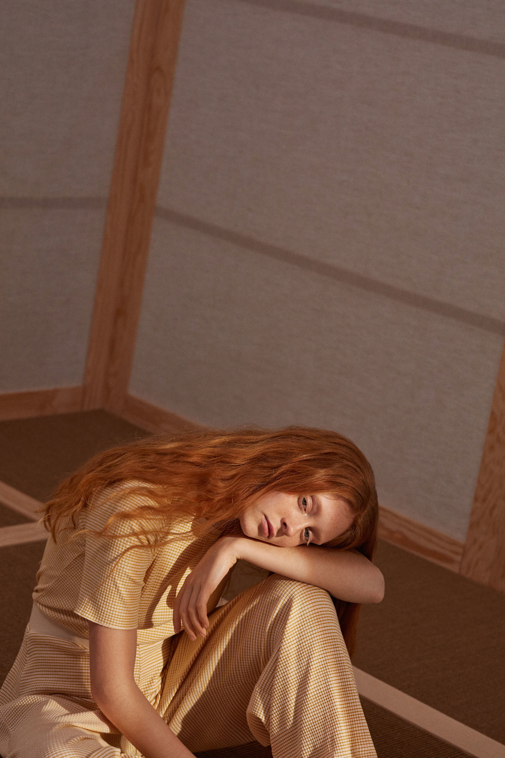 Red-haired woman in a striped outfit sits indoors with her head resting on her knee, bathed in soft, neutral lighting in a minimalist room.