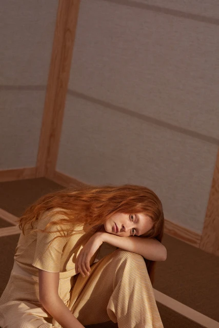 Red-haired woman in a striped outfit sits indoors with her head resting on her knee, bathed in soft, neutral lighting in a minimalist room.