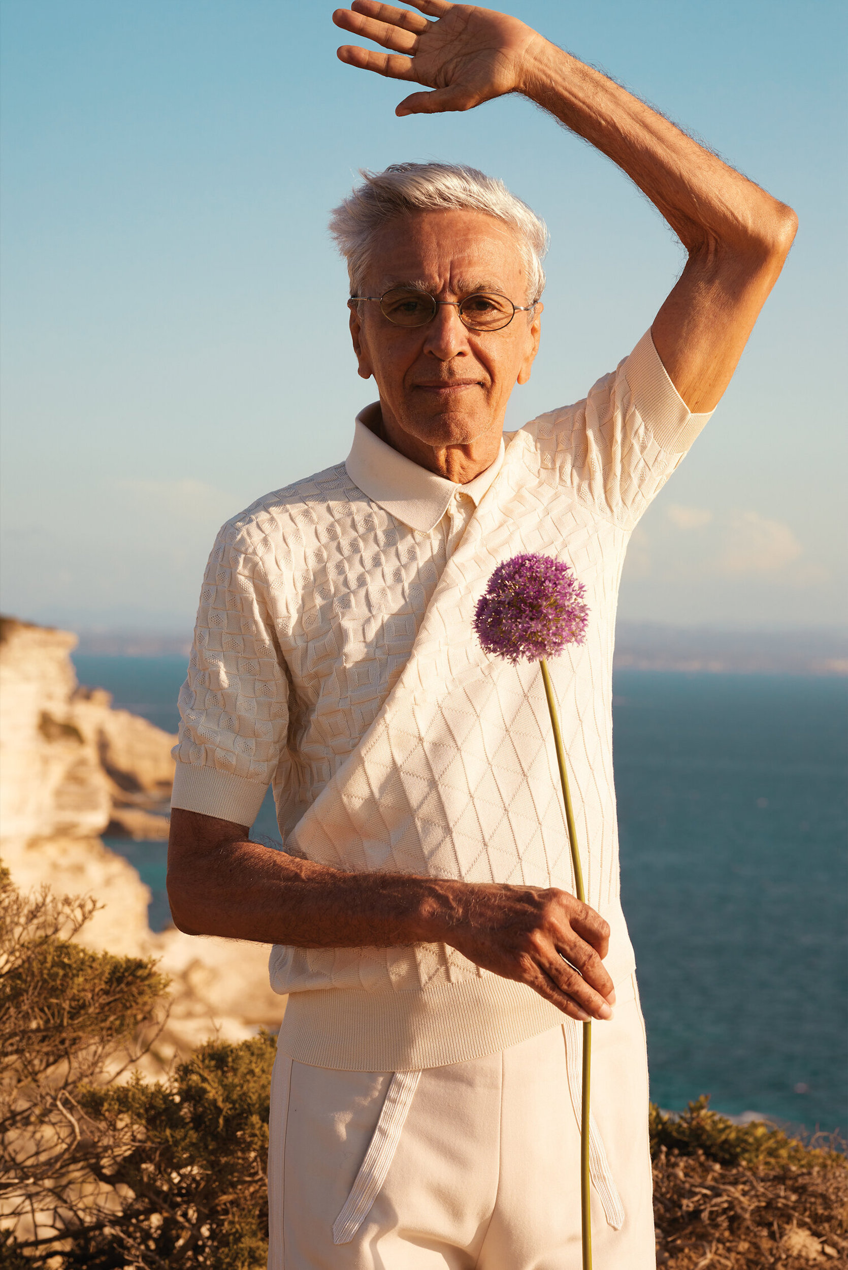 Elderly man in white holds purple allium flower by ocean cliffs under blue sky, wearing a white polo shirt and white pants.