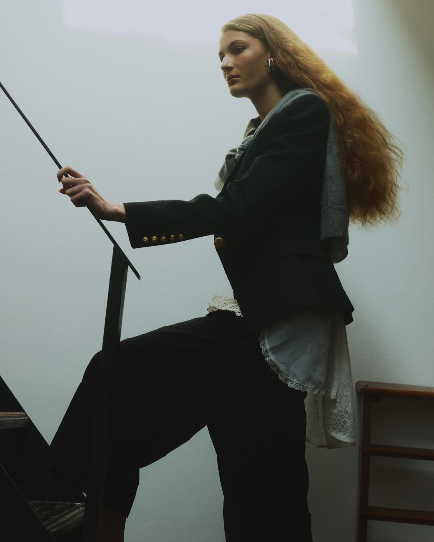 A fair-skinned woman with long wavy hair is ascending stairs while holding onto a black metal railing. She is wearing a dark blazer and pants.