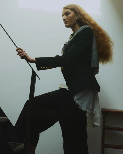 A fair-skinned woman with long wavy hair is ascending stairs while holding onto a black metal railing. She is wearing a dark blazer and pants.