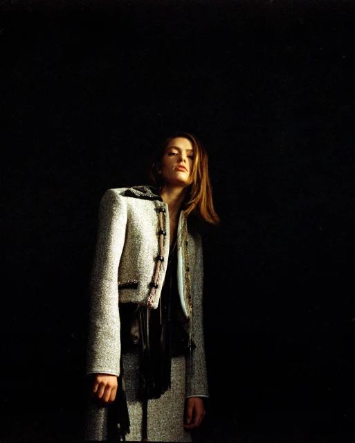 Woman in a gray tweed jacket and skirt posing against a dark background in a studio setting, captured in a fashion photography style.