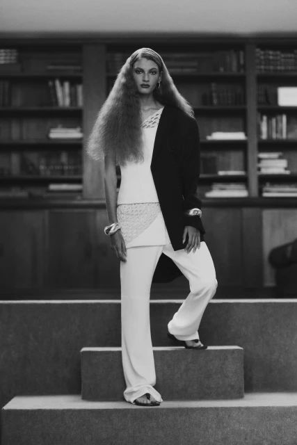 Monochrome studio shot of a woman with long wavy hair wearing a black blazer and white pants stepping down some stairs next to bookshelves.