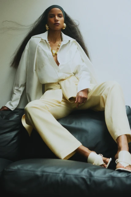 Fashionable Black woman in a cream-colored shirt and pants sits on a leather couch with hair flowing, gold jewelry, and a head scarf.