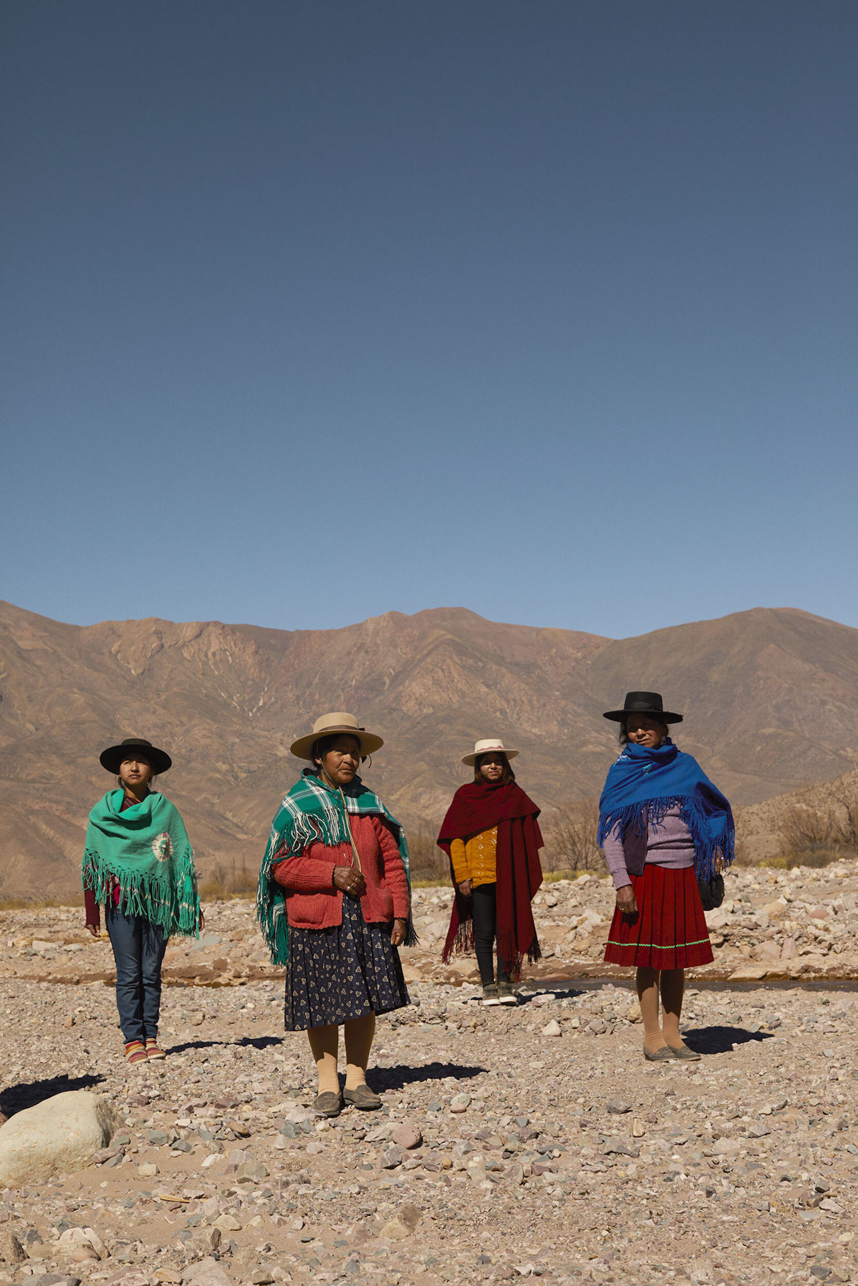 Four women in traditional clothing stand on rocky ground with mountains in the background under a clear blue sky, full shot.