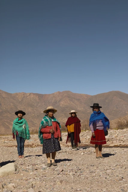 Four women in traditional clothing stand on rocky ground with mountains in the background under a clear blue sky, full shot.
