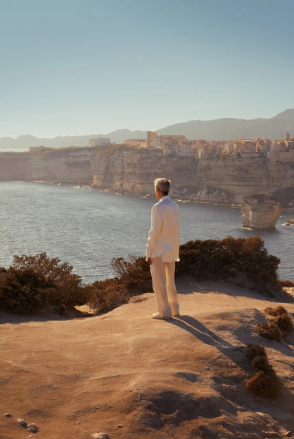 A man in a white suit stands on a rocky cliff overlooking the sea, with a view of coastal cliffs and a distant town on a sunny day.