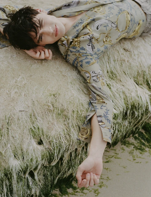 Portrait of a young male model in patterned shirt lying on a mossy stone surface, photographed from a high angle.