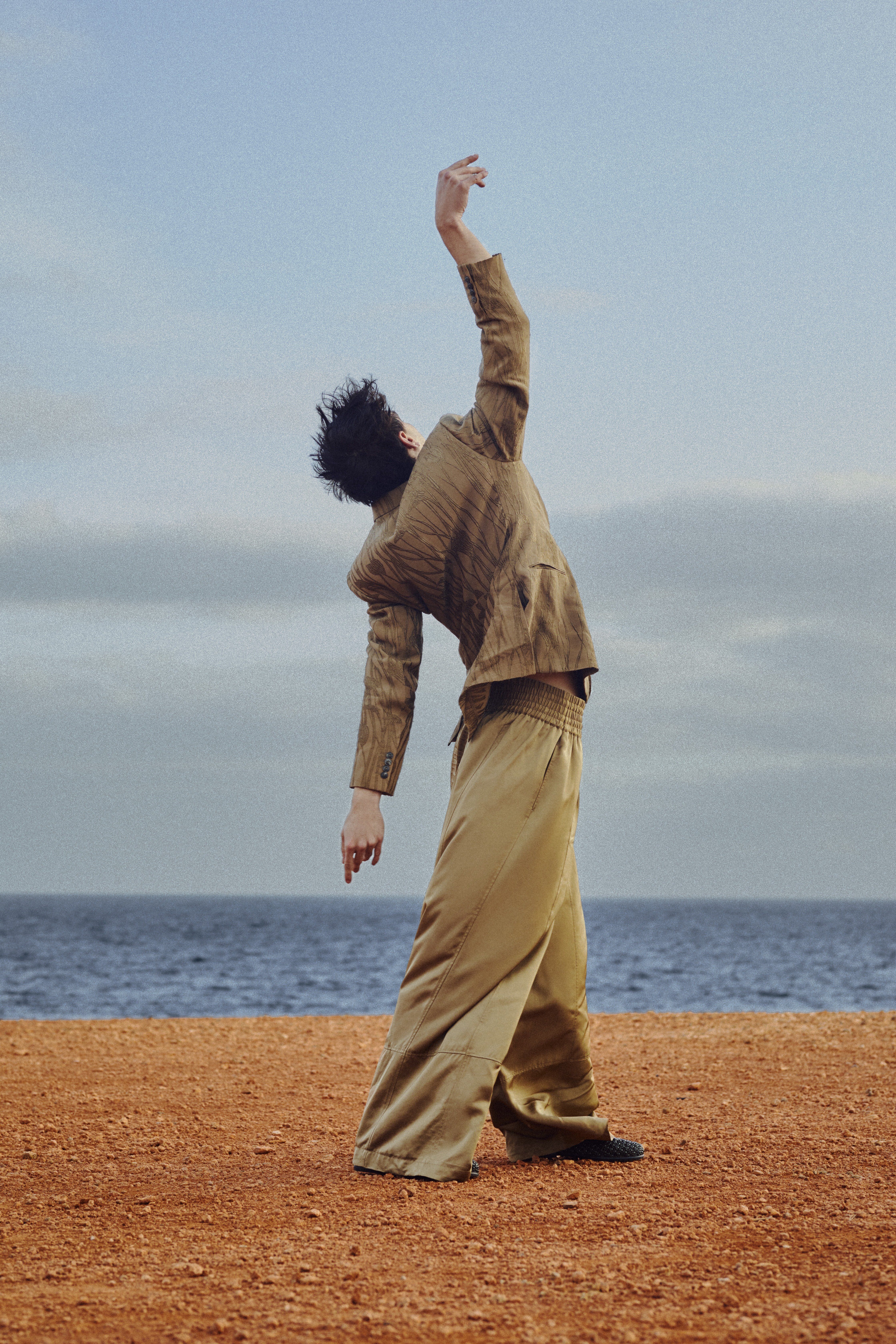 Dancer in beige trousers and a textured jacket posing against an ocean horizon background