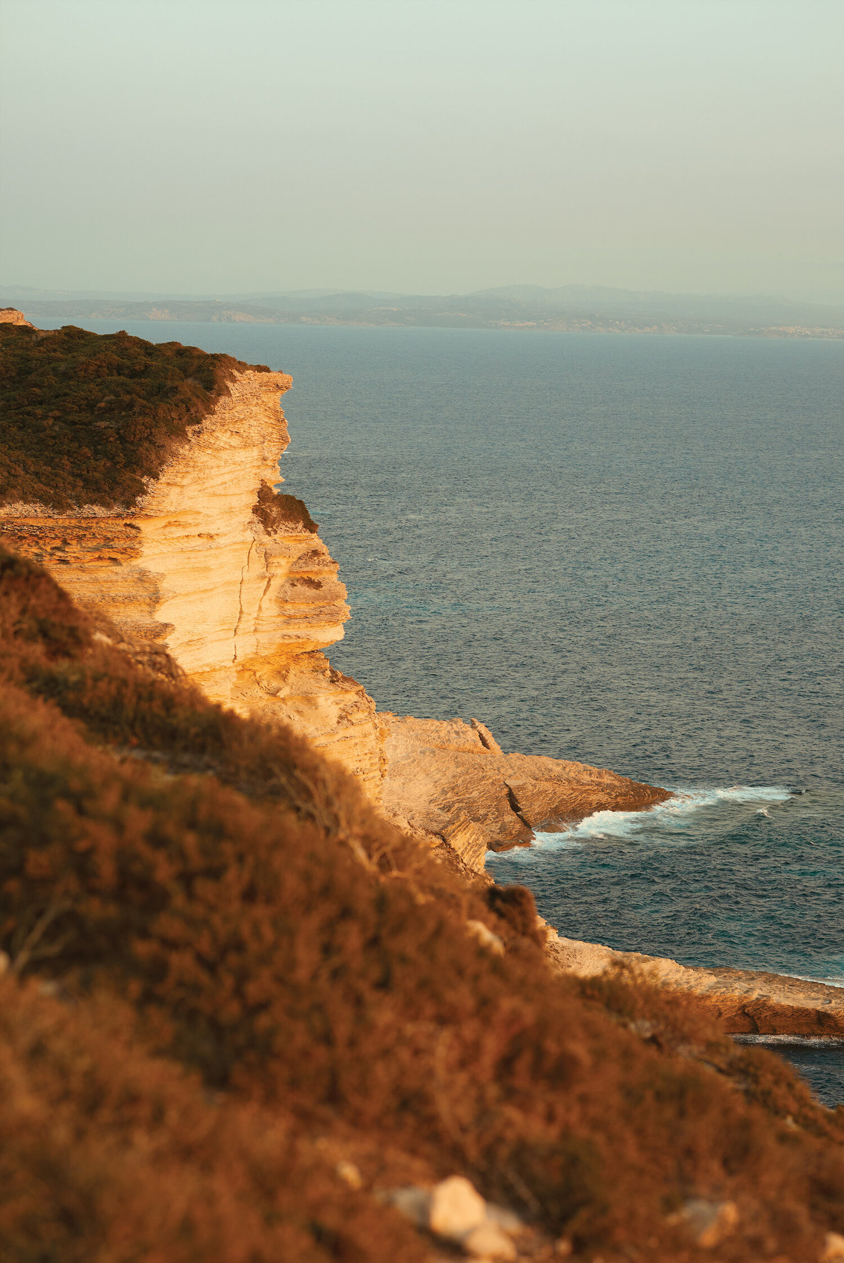 Scenic view of a rocky cliffside with vegetation overlooking the ocean, captured in soft, warm lighting during daytime.